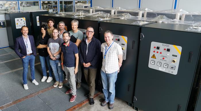 group of people smiling in front of many boxy pieces of equipment with buttons and 'CLIQ3' written on them 